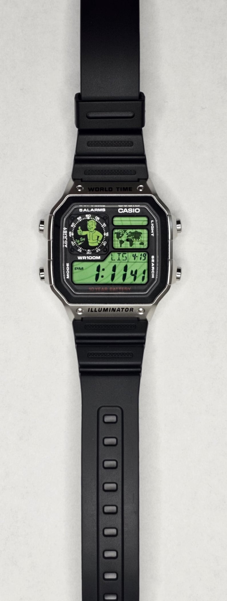 May include: A black digital wristwatch with a silver-tone rectangular case and a black resin strap. The watch face features a green digital display with a world map, the text "WORLD TIME," "CASIO," "ILLUMINATOR," and a graphic of a cartoon character.