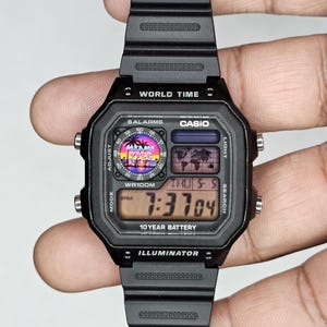 K&ouml;nnte beinhalten: Schwarze Digitaluhr mit rechteckigem Zifferblatt und schwarzem Armband. Das Zifferblatt zeigt die Uhrzeit, den Tag und das Datum an. Die Uhr tr&auml;gt die Aufschrift "WORLD TIME", "CASIO", "10 YEAR BATTERY" und "ILLUMINATOR". Die Uhr tr&auml;gt auch die Aufschrift "MIAMI VICE".
