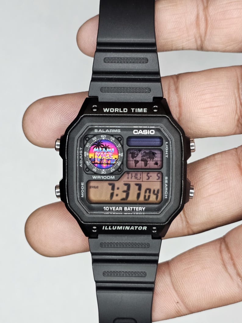 K&ouml;nnte beinhalten: Schwarze Digitaluhr mit rechteckigem Zifferblatt und schwarzem Armband. Das Zifferblatt zeigt die Uhrzeit, den Tag und das Datum an. Die Uhr tr&auml;gt die Aufschrift "WORLD TIME", "CASIO", "10 YEAR BATTERY" und "ILLUMINATOR". Die Uhr tr&auml;gt auch die Aufschrift "MIAMI VICE".