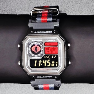 Puede incluir: Un reloj digital Casio negro y plateado con una correa a rayas rojas y negras. La esfera del reloj muestra la hora, un logotipo de Cobra y la palabra "ILLUMINATOR". El reloj está sobre una superficie negra.