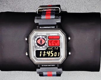Puede incluir: Un reloj digital Casio negro y plateado con una correa a rayas rojas y negras. La esfera del reloj muestra la hora, un logotipo de Cobra y la palabra "ILLUMINATOR". El reloj está sobre una superficie negra.