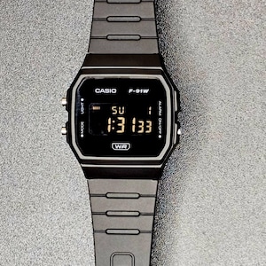 Puede incluir: Reloj digital Casio F-91W negro. El reloj muestra la hora 1:31:33 en una pantalla digital negra. La correa y la caja del reloj son negras. El reloj tiene botones para modo, luz y alarma.