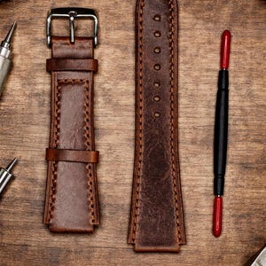 May include: A brown leather watch strap with a silver buckle and contrasting stitching. The strap is shown with a matching extension piece and a watch repair tool on a wooden surface. The strap is approximately 9 inches long.