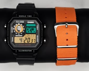 May include: A black digital watch with a portrait on the face and an orange nylon watch strap. The watch face displays the time 4:20:37. The watch is a Casio model.