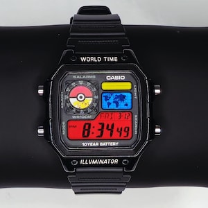 May include: Black digital watch with a rectangular face, displaying the time 8:34:49. The watch face includes the text "WORLD TIME", "CASIO", and "10 YEAR BATTERY". The watch has a black strap and is set on a black surface.