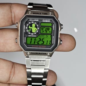 May include: A silver-tone digital wristwatch with a metallic link band. The watch face features a green display with the text WORLD TIME, CASIO, 5 ALARMS, WR100M, UTC, 10 YEAR BATTERY, and ILLUMINATOR. A graphic of a cartoon character is visible.