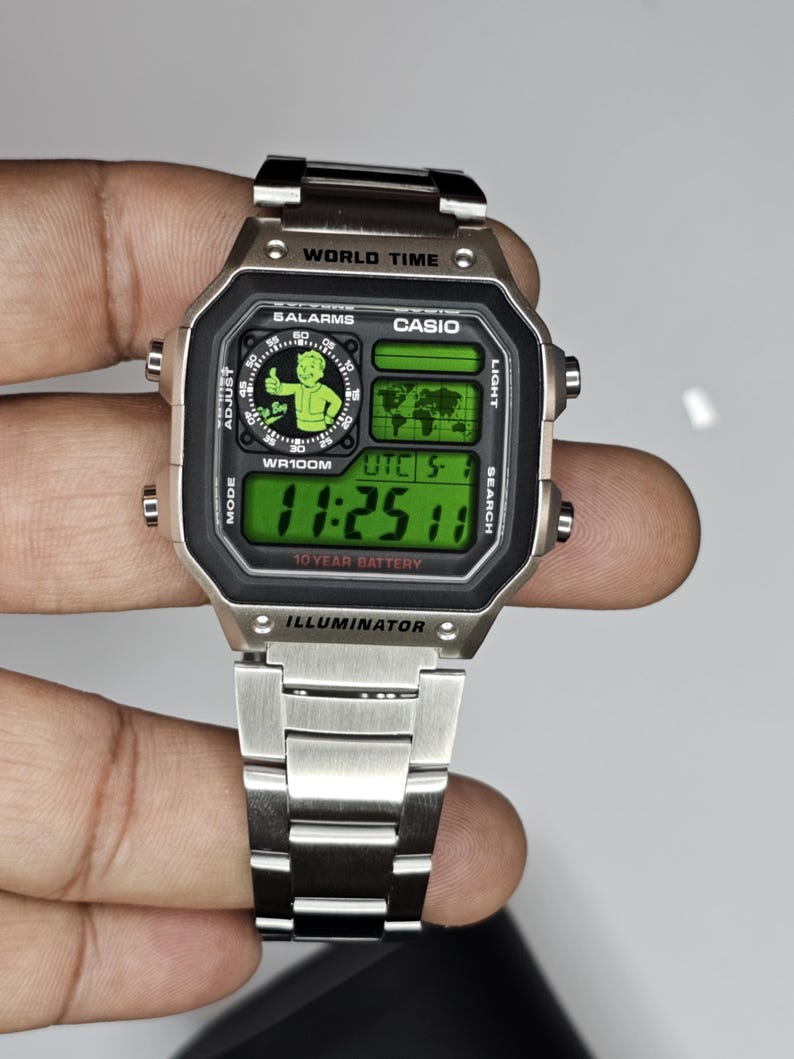 May include: A silver-tone digital wristwatch with a metallic link band. The watch face features a green display with the text WORLD TIME, CASIO, 5 ALARMS, WR100M, UTC, 10 YEAR BATTERY, and ILLUMINATOR. A graphic of a cartoon character is visible.
