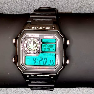 May include: Black Casio digital watch with a green display showing the time 4:20:35. The watch face has a cannabis leaf graphic and a world map. The watch band is black.