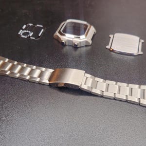 以下が含まれることがあります： 金属バンド、文字盤、裏蓋を備えた、分解されたシルバー調の時計。 時計バンドは留め金を開いた状態で配置されています。 文字盤は暗い画面です。 時計の部品は暗い表面にあります。