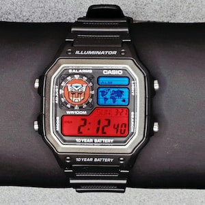 Puede incluir: Reloj digital Casio negro con el logotipo de Transformers en la esfera. El reloj tiene una pantalla digital roja que muestra la hora, la fecha y otras funciones. El reloj tiene una correa negra y está etiquetado como "ILLUMINATOR" y "10 YEAR BATTERY".