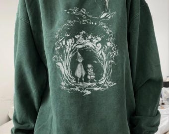 May include: A dark forest green crewneck sweatshirt featuring a white screen-printed graphic of two small figures standing in a wooded clearing beneath a large, dark, antlered creature silhouette looming in the trees above them.