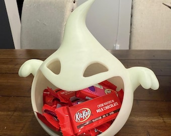 May include: A luminous white ghost-shaped bowl filled with red Kit Kat bars and other wrapped sweets. The bowl features two eye-shaped cutouts and two small arm-like protrusions.