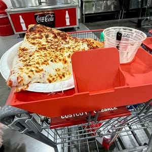 May include: A large slice of pizza on a paper plate, resting in a red shopping cart tray. The pizza has a thick layer of melted cheese and a red sauce base. A clear plastic cup sits next to the pizza.
