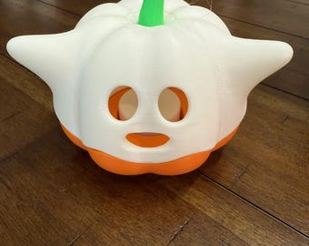 May include: A white and orange pumpkin-shaped decoration with a green stem. The pumpkin has two eye holes and a mouth-shaped opening. The decoration is sitting on a wooden surface.