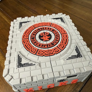 May include: A gray, rectangular puzzle box with a brick-like texture. The top features an orange and black circular design. The word "KICKS" is in orange letters on the side. The box is on a wooden surface.