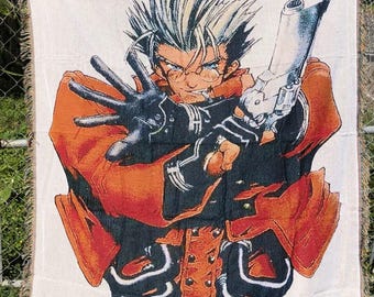 Può includere: Una coperta tessuta di Trigun Maximum con un'illustrazione di un personaggio con una pistola. Il personaggio ha i capelli biondi a spillo, occhiali e una giacca arancione. La coperta ha uno sfondo bianco con il testo "TRIGUN MAXIMUM" in alto.