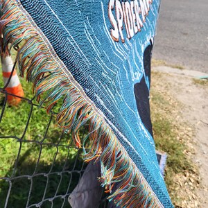 May include: A blue woven throw blanket featuring the text "THE AMAZING SPIDER-MAN" in white and orange. The blanket has a fringed edge with orange, green, and blue threads. The blanket is made of a woven material.