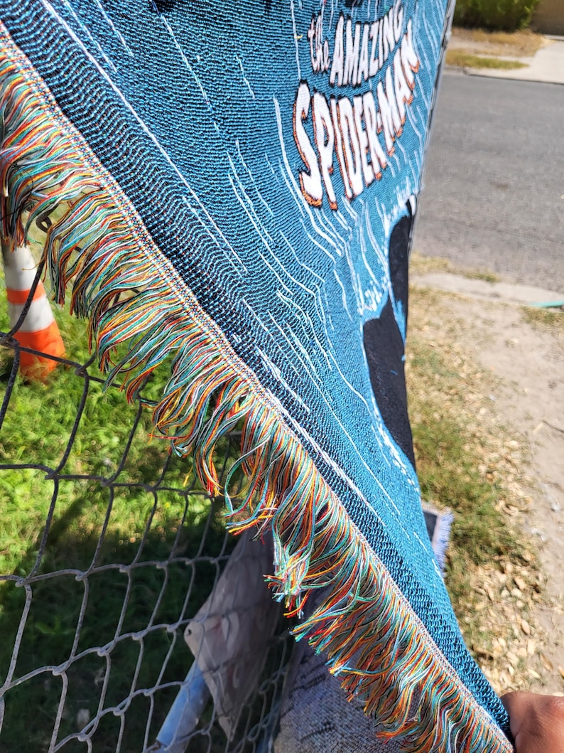 May include: A blue woven throw blanket featuring the text "THE AMAZING SPIDER-MAN" in white and orange. The blanket has a fringed edge with orange, green, and blue threads. The blanket is made of a woven material.