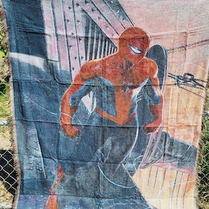 May include: A woven throw blanket featuring a vibrant illustration of Spider-Man in a dynamic pose. The blanket showcases the superhero in his iconic red and blue suit against a backdrop of a cityscape. The blanket has a fringed edge.