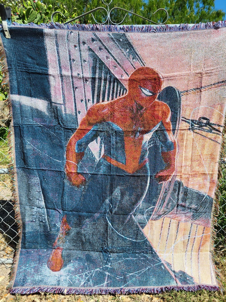 May include: A woven throw blanket featuring a vibrant illustration of Spider-Man in a dynamic pose. The blanket showcases the superhero in his iconic red and blue suit against a backdrop of a cityscape. The blanket has a fringed edge.