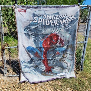 May include: A woven throw blanket featuring the Amazing Spider-Man. The blanket displays the Marvel logo and the words "The Amazing Spider-Man" with an image of Spider-Man in his red and blue suit. The blanket is hanging on a fence.