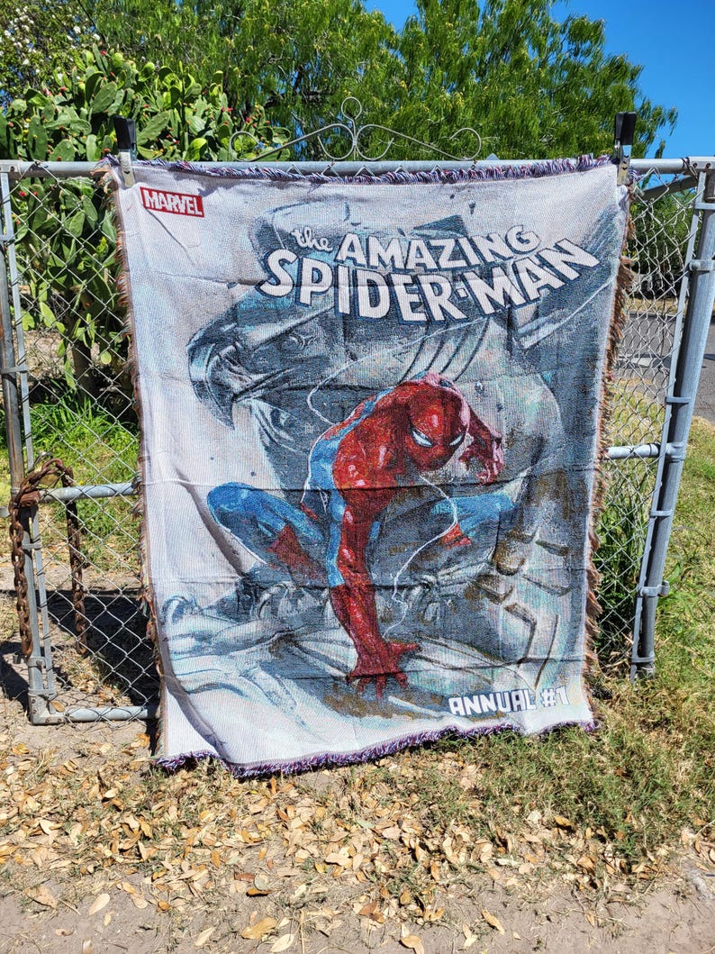 May include: A woven throw blanket featuring the Amazing Spider-Man. The blanket displays the Marvel logo and the words "The Amazing Spider-Man" with an image of Spider-Man in his red and blue suit. The blanket is hanging on a fence.