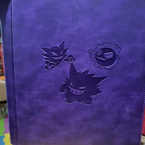 May include: A rectangular purple binder featuring embossed illustrations of three distinct ghost-type creatures on the front cover. The surface has a smooth, matte texture with a vertical stitched seam running along the left side of the cover.