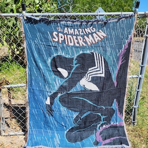 May include: A blue woven throw blanket featuring the Amazing Spider-Man comic book cover art. The design shows Spider-Man in a black suit, kneeling in the rain. The blanket has a fringed edge and is displayed outdoors.