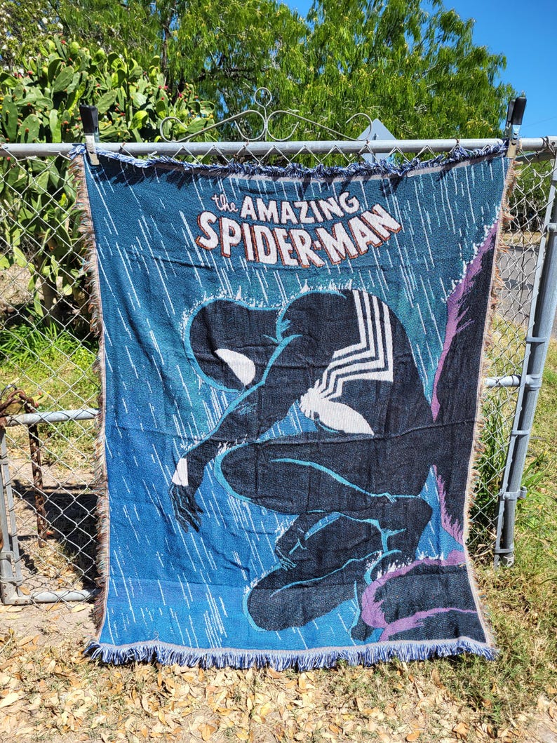 May include: A blue woven throw blanket featuring the Amazing Spider-Man comic book cover art. The design shows Spider-Man in a black suit, kneeling in the rain. The blanket has a fringed edge and is displayed outdoors.