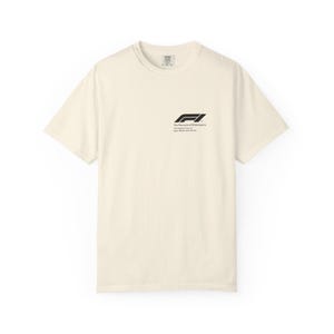 Puede incluir: Camiseta de manga corta color crema con el logotipo de Fórmula 1 y texto. El logotipo negro presenta una "F" y un "1" estilizados. El texto de abajo dice "The Pinnacle of Motorsports. The Highest Class of Open-Wheel Auto Racing."
