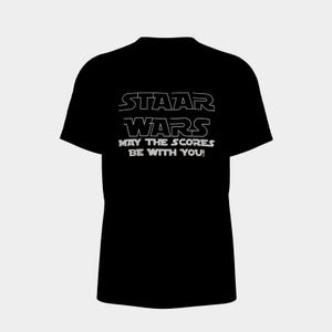 May include: Black t-shirt with white text that reads "STAAAR WARS" above "MAY THE SCORES BE WITH YOU!". The shirt has a crew neck and short sleeves. The design is a parody of the Star Wars logo.