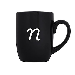 May include: A black ceramic mug with a white cursive letter "n" on the front. The mug has a curved handle and a smooth, glossy finish. The mug is designed for hot or cold beverages.