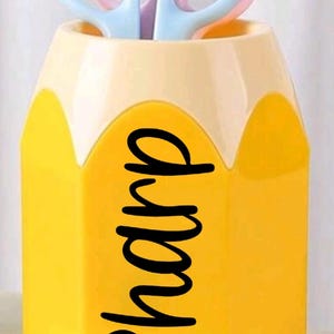 Puede incluir: Contenedor amarillo con forma de lápiz con la palabra "sharp" en escritura negra. El contenedor contiene dos pares de tijeras, uno rosa y otro azul. La parte superior del contenedor es de color amarillo claro.