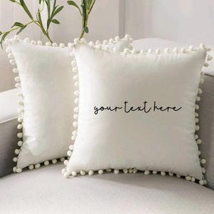 Könnte beinhalten: Zwei quadratische, cremefarbene Zierkissen mit weicher Textur und einem Rand aus kleinen, weißen Pompons. Ein Kissen hat den Text "your text here" in handschriftlichem Stil. Die Kissen liegen auf einem hellgrauen Sofa.