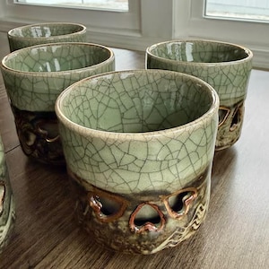 May include: A set of four ceramic cups with a unique crackle glaze in shades of green and brown. The cups have a distinctive design with cut-out shapes near the base, adding a touch of artistry to the functional drinkware.