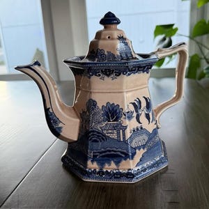 May include: A vintage ceramic teapot with a blue and white Willow pattern design. The teapot has a beige body, a blue lid, and a curved spout. The handle is also beige. The design features a landscape scene with bridges and trees.