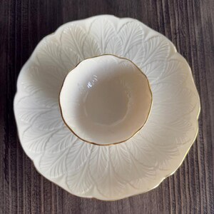 May include: A cream-colored, decorative serving set featuring a large, leaf-patterned plate and a smaller bowl. The bowl and plate have gold trim. The set is designed for serving food or displaying decorative items.