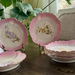 May include: A set of vintage porcelain dishes with a pink scalloped edge and gold trim. The plates and bowls feature floral designs in the center. The set includes two plates and a stack of bowls.