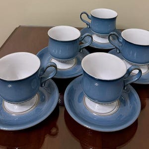 May include: A set of six blue and white teacups and saucers. The cups have a rounded shape with a handle, and the saucers are round. The set is made of ceramic and has a glossy finish. The cups and saucers are arranged on a dark brown surface.