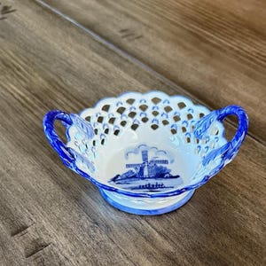May include: A small, white and blue ceramic dish with two handles. The dish has a scalloped edge and a windmill design in the center. The handles and rim are blue. The dish is sitting on a wooden surface.