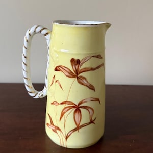 May include: A yellow ceramic pitcher with a white and gold handle. The pitcher is decorated with brown floral designs. The rim of the pitcher is trimmed with gold. The pitcher is sitting on a dark brown surface.