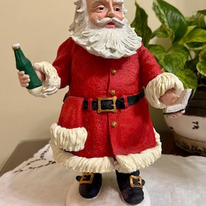 May include: A Santa Claus figurine wearing a red coat with white fur trim, a black belt with a gold buckle, and black boots. The figure has a white beard, gold-rimmed glasses, and holds a green glass bottle in one hand and an open book in the other.