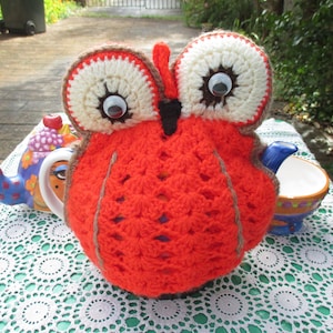 Peut inclure: Une housse de théière en forme de hibou en crochet orange avec des accents marrons et de grands yeux blancs. La housse est posée sur une nappe en crochet blanche.