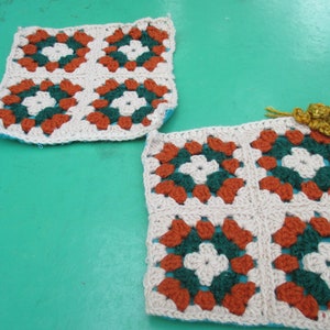 May include: Two handmade crochet squares with a floral pattern. Each square features a beige background with orange, green, and white floral designs. The squares are placed on a teal surface, showcasing the intricate details of the yarn work.