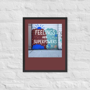 Puede incluir: Una impresión de arte mural enmarcada que presenta un mural con el texto FEELINGS ARE SUPERPOWERS en letras mayúsculas blancas. El mural muestra un diseño dividido con un lado rojo oscuro y otro verde azulado, decorado con iconos azules.