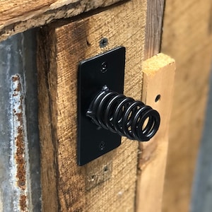 May include: A black metal door latch with a spring attached to a wooden door frame. The door frame is made of reclaimed wood with a rustic finish. The door is made of corrugated metal with rust and patina.