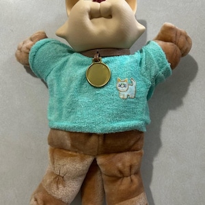 May include: A plush cat doll with a molded vinyl head, brown yarn hair, and brown fabric limbs. It wears a teal short-sleeved shirt featuring an embroidered cat graphic and a brown collar with a round gold-colored metal tag hanging at the center.