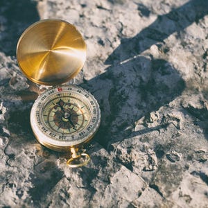 May include: A vintage brass compass with a gold-colored lid, resting on a textured, gray stone surface. The compass face features directional markings and a needle. The lid is open, casting a shadow.