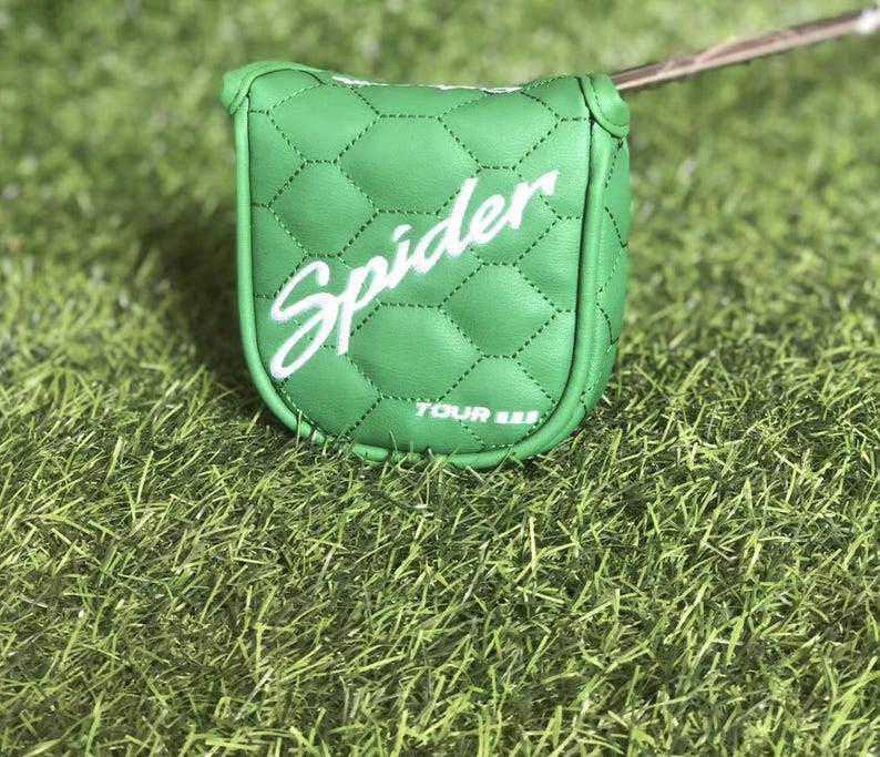 Puede incluir: Una funda para la cabeza del putter de golf verde con la palabra "Spider" en escritura blanca. La funda tiene un patr&oacute;n acolchado y el texto "TOUR III" en la parte inferior. La funda descansa sobre un lecho de hierba verde.