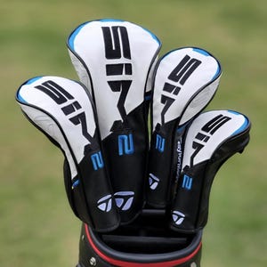 May include: A collection of four golf club head covers in a golf bag. The covers are white with black and blue detailing, displaying the number "2" and the letters "SIF" in black. The TaylorMade logo is visible.